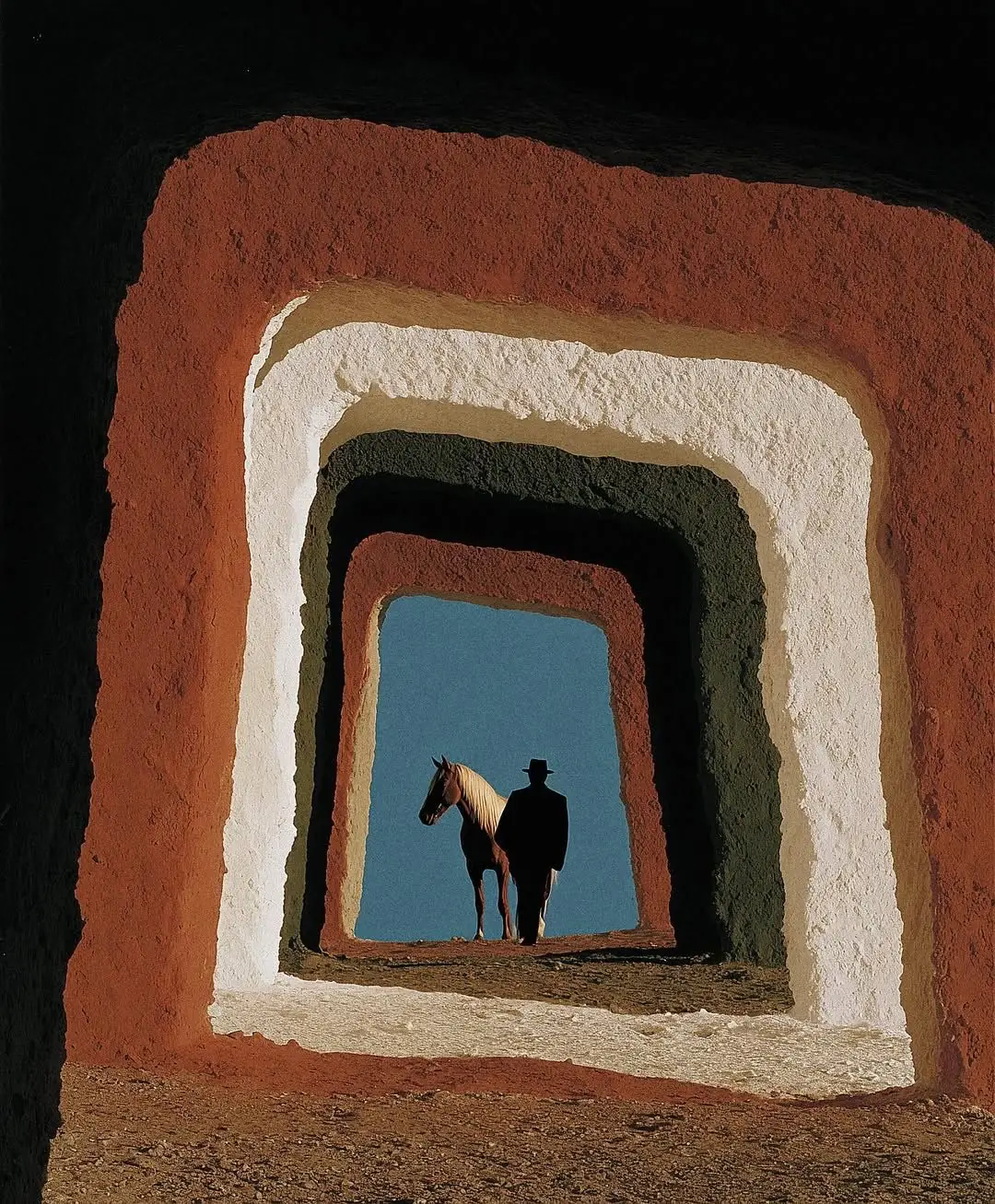 Une porte de terre rouge et blanche, puis une autre, et une autre ; tout au fond, un homme et son cheval devant le ciel.
